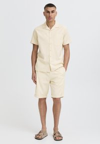 Camicia gialla chiaro a maniche corte con collo e taschino abbinata a pantaloncini coordinati, caratterizzati da una vestibilità rilassata e orli arrotolati. Sandali marroni.