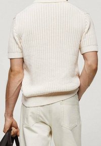 Polo de punto de manga corta en color crema con un patrón de canalé texturizado, que presenta un cuello y un corte relajado, combinado con pantalones de color claro.