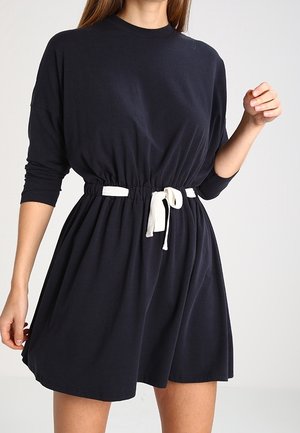 Kobieta ubrana w czarną sukienkę z długim rękawem z białym troczkiem w talii, stojąca na tle jednolitego jasnego tła.