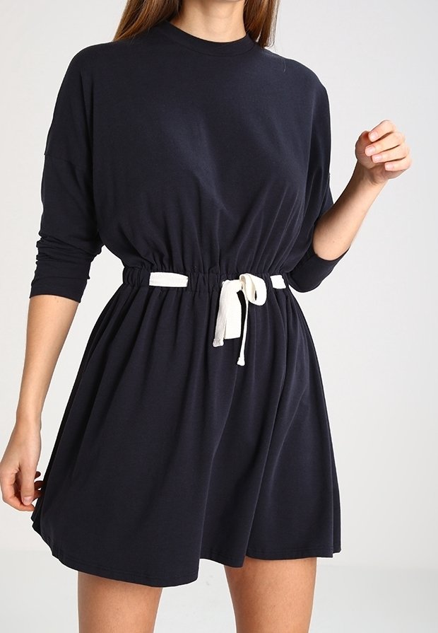 Femme portant une robe noire à manches longues avec une ceinture à cordon blanc à la taille, debout devant un arrière-plan clair uni.