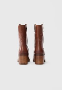 Un paio di stivali ankle in pelle marrone con tacco in legno, mostrati dal retro su uno sfondo bianco semplice.