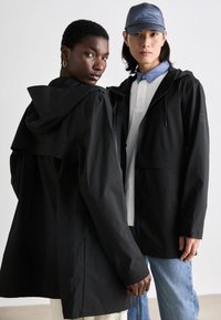 Jachete negre din material ușor, cu glugă, având o croială lejeră, textură netedă și un design minimalist. Un model poartă o cămașă albastră deschisă dedesubt.