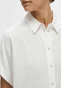 Chemise blanche à col pointu avec une texture douce, manches courtes et motif subtil. Elle se distingue par de petits boutons.