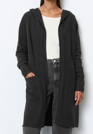 Femme portant un long cardigan à capuche foncé sur un top blanc côtelé et un jean noir délavé taille haute, une main dans la poche.