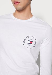 T-shirt bianco a maniche lunghe con un logo circolare che presenta il testo "TOMMY HILFIGER", la data di fondazione e un design a blocchi di colore in rosso, bianco e blu.