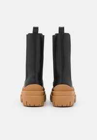 Zwarte vegan leren enkelboots met een bruine chunky zool, met een gladde textuur, ronde neus en een treklus aan de achterkant.