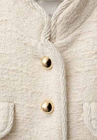 Chaqueta blanca texturizada con una tela tejida, botones de hardware dorados, detalles de cuerda trenzada en el cuello y bolsillos con solapa.
