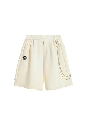 Joven con rastas lleva una camiseta azul marino oversize con el logo "MWM", pantalones cortos blancos, calcetines altos blancos y zapatos beige con textura sin cordones.