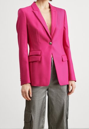 Kobieta ubrana w jasnoróżową dopasowaną marynarkę z patkami i srebrnym guzikiem, zestawioną z luźnymi metalicznymi szarymi spodniami typu cargo.