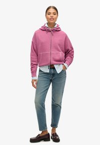 Sweatshirt à capuche fuchsia avec poches avant, fermeture éclair et cordons de serrage. Associé à une chemise bleu clair, un jean bleu et des mocassins bourgogne.