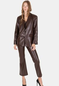 Donna che indossa un blazer in pelle marrone scuro e pantaloni cropped abbinati, abbinati a tacchi alti neri, in piedi contro uno sfondo semplice.
