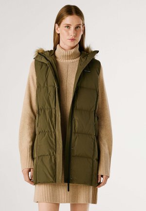 Gilet imbottito verde oliva con cappuccio, chiusura con zip e rifinitura in pelliccia. Indossato sopra un dolcevita beige a costine e un abito. Presenta tasche laterali.