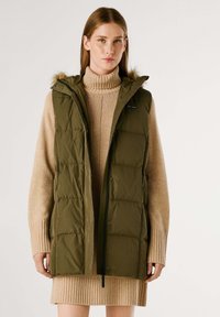 Padded oliwkowa kamizelka z kapturem, z zamkiem z przodu i klapami. Noszona na beżowym swetrze z golfem w prążki.