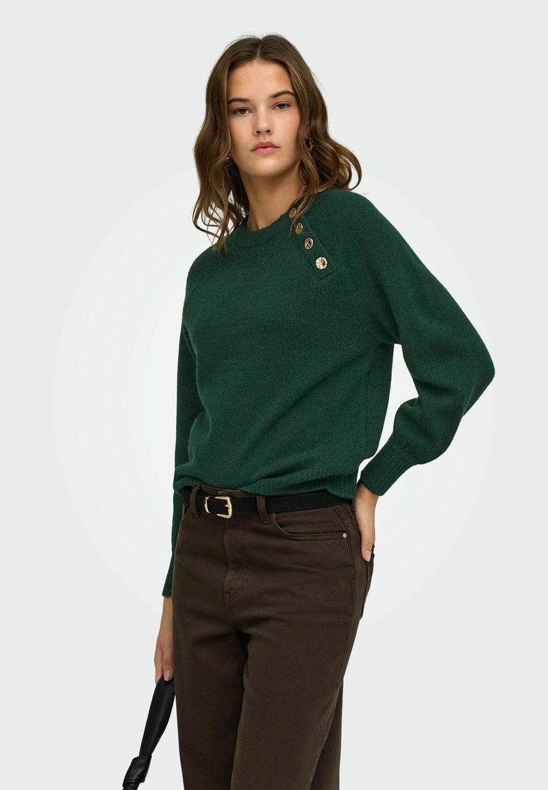 Femme portant un pull vert foncé avec des boutons dorés sur l'épaule, un pantalon marron foncé, et tenant un sac à main noir.