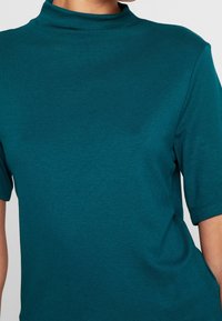 Teal kortærmet top med høj hals. Stoffet ser blødt ud med en subtil tekstur og afslappet pasform. Ingen synlige mønstre eller detaljer.