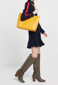 Żółta skórzana torba typu tote o wyraźnej formie, z metalowymi złotymi elementami oraz długim łańcuszkowym paskiem. Nosi się ją z granatową sukienką i brązowymi kozakiem za kolano.