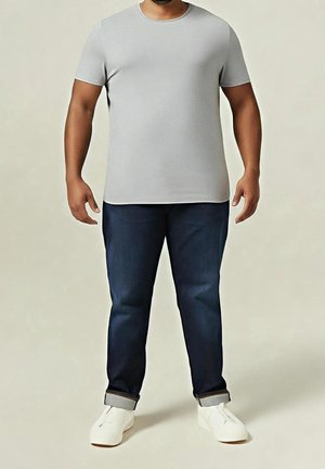 Man som bär en ljusgrå t-shirt med kort ärm, mörkblå jeans med uppvikta fållar och vita skor, stående mot en neutral bakgrund.