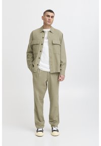 Oliivinvihreä, kevyt kaksiosainen asu, johon kuuluu napillinen paita, jossa on kaksi rintataskua, ja siihen sopivat housut. Asu on yhdistetty mustavalkoisiin tennareihin.