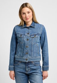 Veste en denim bleu clair au design ajusté, boutons dorés, deux poches poitrine et un col classique. Détails de couture visibles.