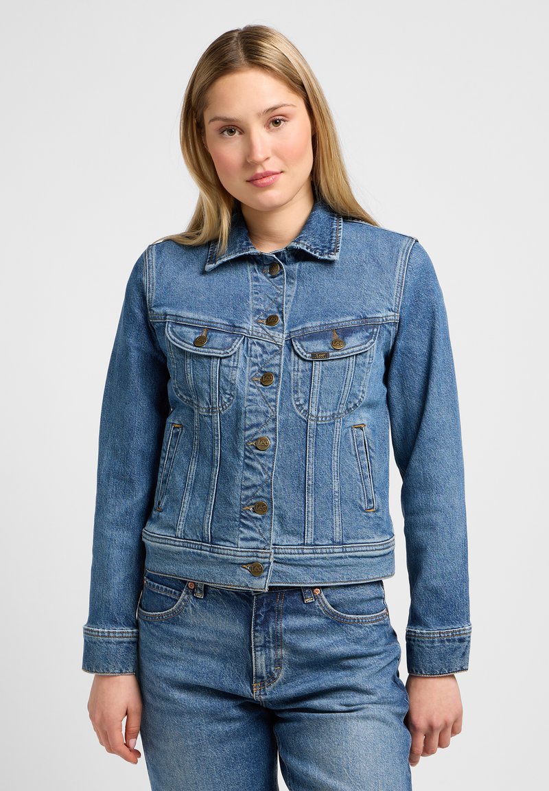 Veste en denim bleu clair au design ajusté, boutons dorés, deux poches poitrine et un col classique. Détails de couture visibles.