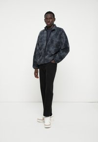 Fleece-zipperjacke in Dunkelgrau mit weichem Material, getragen über einem schwarzen Shirt und kombiniert mit schwarzen Hosen und weißen Sneakern.