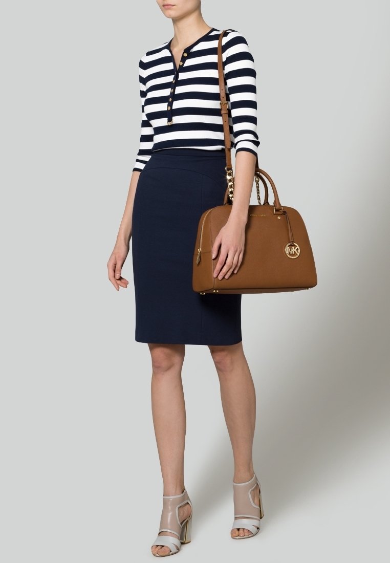 Femme en jupe crayon bleu marine et haut à manches longues rayé noir et blanc, tenant un sac à main en cuir marron avec des accents dorés, portant des talons beiges ouverts.