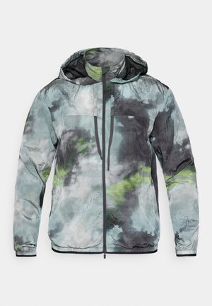Chaqueta ligera en tonos de verde, gris y negro. Cuenta con capucha, cierre frontal y una tela texturizada con patrones abstractos.