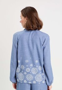 Chemise bleu à manches longues en tissu léger, dotée d'une broderie florale blanche dans le dos, d'une finition texturée et de poignets élastiques.