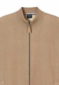 Veste beige à fermeture éclair avec un matériau côtelé texturé, col montant et accents métalliques subtils le long de la fermeture éclair et des bords des poches.