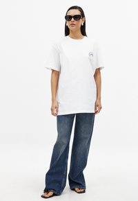 T-shirt en coton blanc à manches courtes avec un petit motif bleu, assortie à un jean en denim bleu foncé évasé et des lunettes de soleil noires.