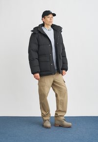 Fekete szigetelt puffertöltetű dzseki gallérral, elülső zsebekkel; világoskék fonott mintás pulóverrel és bézs cargo nadrággal párosítva. A sötét cipő egészíti ki a ruhát.