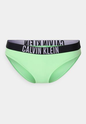 Zaļa bikini apakša, izgatavota no gludas auduma, ar melnu un pelēku elastīgu jostasvietu, uz kuras ir zīmola nosaukums "Calvin Klein."