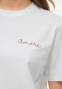 Λευκό βαμβακερό T-shirt με κόκκινη κεντημένη λέξη «Amore» με καλλιγραφική γραφή στο αριστερό στήθος, φορεμένο από ένα άτομο με ανοιχτόχρωμο δέρμα και μακριά μαλλιά.