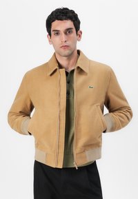 Veste bomber beige avec une fermeture éclair à l'avant, des poignet et un ourlet côtelés. Présente un logo vert et est portée par-dessus un polo vert olive.