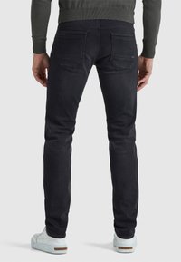 Uomo che indossa jeans slim-fit scuri e sneaker bianche, visto da dietro su uno sfondo grigio uniforme.
