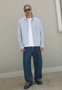 Camicia a righe blu chiaro con bottoni sopra una t-shirt bianca, abbinata a jeans scuri larghi e scarpe nere massicce con lacci.