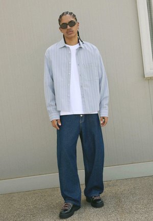 Hellblau gestreifte Button-Up-Bluse über einem weißen T-Shirt, kombiniert mit weiten dunkelblauen Jeans und klobigen schwarzen Schnürschuhen.