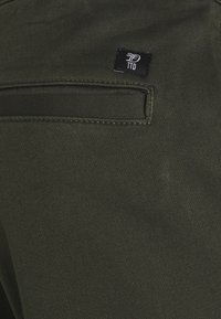 Olivgröna byxor med en slät yta, med en bakficka och en liten svart logotyp sydd på tyget.