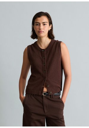 Frau mit kurzem dunklem Haar, die eine ärmellose braune gestrickte Weste mit Knöpfen und passende braune Hose trägt, mit den Händen in den Taschen.
