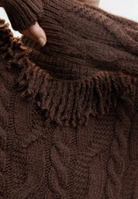 Pull en tricot marron avec un motif torsadé texturé et un col à froufrous. Manches longues avec design sans doigts. Matière douce et épaisse.