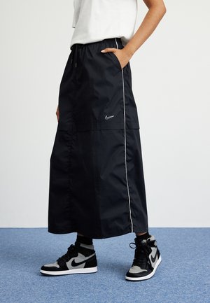 Personne portant une longue jupe noire Nike avec un liseré blanc et des baskets montantes Nike noires et grises sur un sol recouvert de moquette bleue.