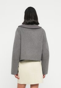 Γκρι cropped μάλλινο σακάκι με φαρδιά ριμπ λαιμόκοψη και μακριά μανίκια, με υφασμάτινη υφή. Φοριέται πάνω από ανοιχτόχρωμη δερμάτινη φούστα.