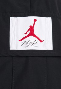 Silhouette rouge d'un joueur de basket sautant avec un ballon au-dessus de la tête sur un patch blanc avec un texte noir "Flight" sur un tissu noir.