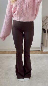 Osoba oblečená v růžovém pleteném svetru a tmavě hnědých rozšířených kalhotách stojící na kobercové podlaze v bílých ponožkách.