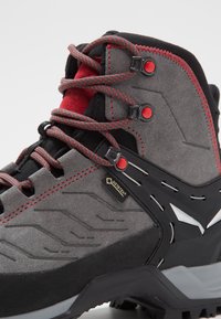 Harmaa vaelluskengä, jossa on mustia ja punaisia yksityiskohtia, valmistettu mokasta ja synteettisestä materiaalista, teksturoidulla pinnalla ja GORE-TEX® -brändillä.