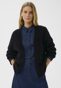 Schwarze Strickjacke mit übergroßen Ärmeln, offene Vorderseite, getragen über einem blauen Denimhemd. Weiche Textur, lässiges Design und starker Farbkontrast.