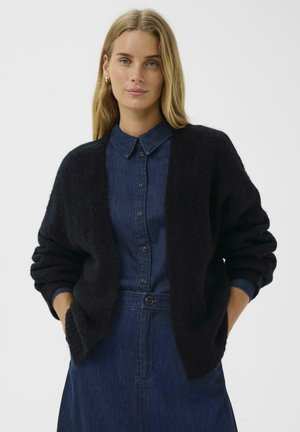 Cardigan noir en tricot avec des manches oversize, à ouverture devant, porté sur une chemise en denim bleu. Texture douce, design décontracté, et fort contraste de couleur.