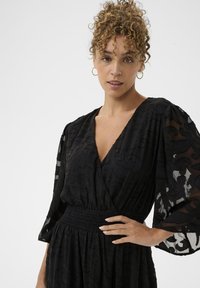 Fekete ruha áttetsző, mintás anyaggal, V alakú nyakkivágással, bő ujjal és húzott derékkal, amely jól meghatározott sziluettet alkot.