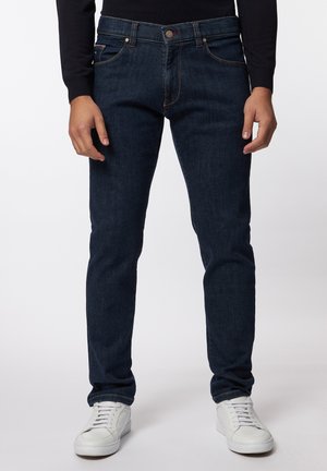 Mand iført mørkeblå slim-fit jeans og hvide sneakers, stående mod en ensartet lys baggrund.