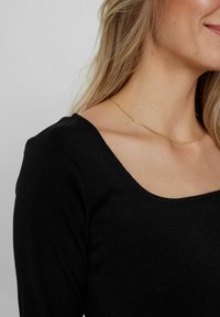Černý žebrovaný top s dlouhým rukávem a čtvercovým výstřihem, s hladkým povrchem. Na krku je viditelný tenký zlatý řetízek.
